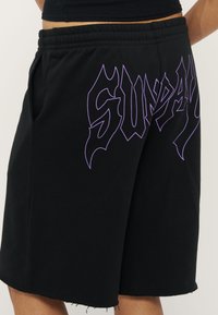 Shorts taille élastique noire avec ourlet effiloché et grand texte violet contouré indiquant "SUNDAY" sur le devant.
