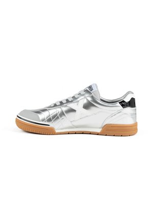 Zapatilla plateada de perfil bajo con cordones blancos, suela de goma, diseño lateral en X cosido y detalle negro en el talón sobre fondo blanco.