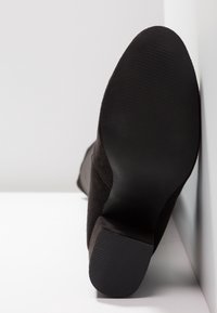 Botte en daim noir à talon bloc bas, vue de la semelle, appuyée contre un mur blanc.