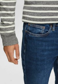 Pull rayé gris et blanc avec poignets côtelés, associé à un jean en denim bleu foncé avec une poche avant et des accents de coutures visibles.