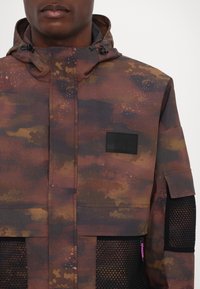 Veste avec motif camouflage marron et orange foncé, poches en mesh noir à l'avant et sur la manche, et écusson noir sur la poitrine.
