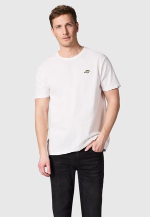 Man draagt een wit t-shirt met korte mouwen met klein planeetlogo op de borst en zwarte broek, staand tegen een effen grijze achtergrond.