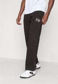 Man wearing black sweatpants with "City" text on thigh, white athletic shoes, and a white shirt against a plain background.