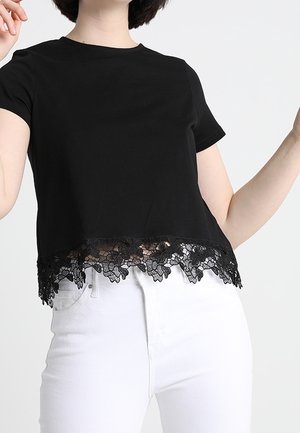 Zwart cropped t-shirt gemaakt van zachte stof, met een kanten randje aan de zoom met bloemmotieven. Draagt wit broek.