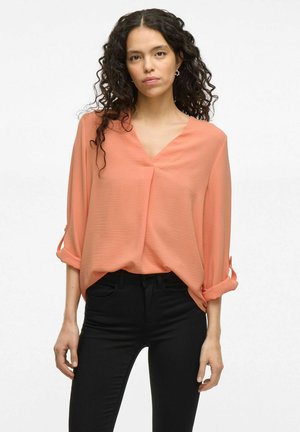 Blouse de couleur pêche, ample, avec un décolleté en V et des manches retroussables, fabriquée en tissu léger, associée à un jean noir ajusté.