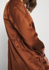 Brun satinklänning med en samlad midja, långa ärmar och en slät textur. Tyget har en subtil lyster som framhäver den eleganta draperingen.