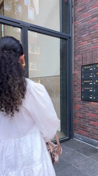 Abito bianco con texture e maniche a sbuffo, borsa di colore chiaro, porta in vetro moderna, edificio in mattoni rossi e marroni, cassette postali sulla parete.