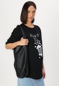 Juoda marškinėliai su užrašu „Ready to Pop“, dekoruoti spragėsių grafika. Suderinti su dideliu juodu tekstūruotu rankiniu krepšiu. Matomos mėlynos džinsinės kelnės.