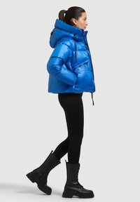 Sijajna modra puhasta jakna z visokim ovratnikom, sprednjo zadrgo in stranskimi žepi. Oblečena s črnimi legicami in robustnimi črnimi škornji.