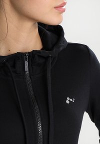 Femme portant un sweat à capuche noir zippé avec cordons et un petit logo réfléchissant sur la poitrine, montrée du cou à l'épaule.