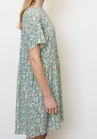 Žena v volných, krátkých šatech s pastelovým květinovým vzorem stojící před jednoduchým bílým pozadím.