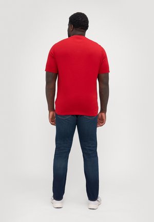 Mann trägt ein schlichtes rotes T-Shirt mit kurzen Ärmeln, dunkelblaue Jeans und weiße Turnschuhe, steht mit dem Rücken zur Kamera auf einem schlichten weißen Hintergrund.