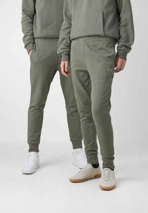 Twee mensen dragen olijfgroene joggerbroeken en bijpassende hoodies met witte en beige sneakers op een effen achtergrond.