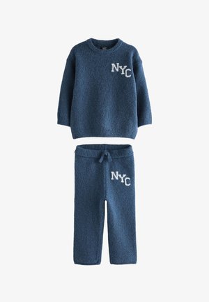 Jedes junge Kind steht in einem blauen Strickpullover und einer Hose mit dem Aufdruck "NY" und trägt weiße Turnschuhe vor einem beigen Hintergrund.