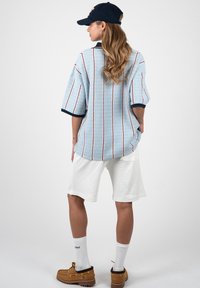 Femme portant une chemise rayée bleu clair, un short blanc longueur genou, des chaussures bateau marron, des chaussettes blanches et une casquette sombre, debout de dos.