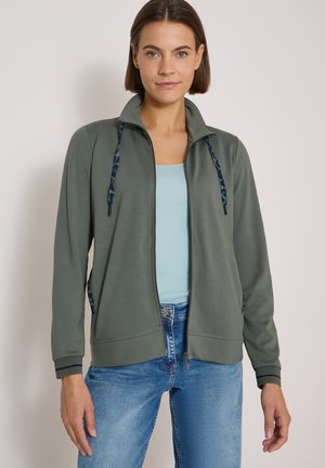 Donna con capelli castani corti indossa una giacca verde con cerniera sopra una maglietta azzurro chiaro e jeans blu, in piedi davanti a uno sfondo chiaro uniforme.
