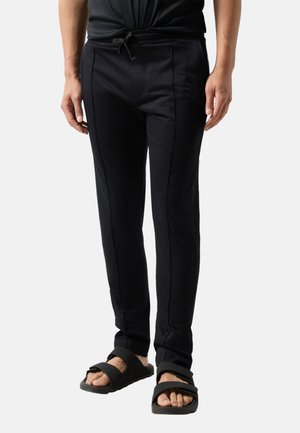 Mężczyzna w czarnych dopasowanych spodniach z troczkiem w pasie i czarnych sandałach z otwartym palcem, stojący na jednolitym tle.