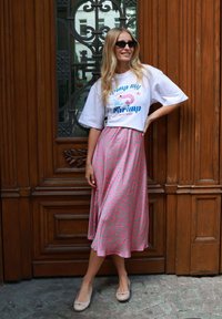 Smiling woman with long blonde hair wears sunglasses, white graphic tee, pink patterned skirt, and beige flats, standing by ornate wooden door.