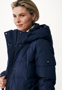 Giacca imbottita blu navy con colletto alto, chiusura con zip e accenti. Presenta un design elegante con un sottile logo del marchio sulla manica.