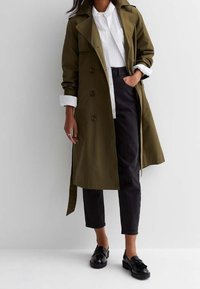 Kvinna iklädd en olivgrön trenchcoat över en vit skjorta och svarta cropped byxor med svarta blanka loafers, stående mot en vit vägg.