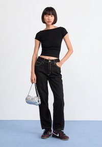 Top court noir côtelé, jeans noir à jambe large avec des coutures blanches, baskets rouges et noires, et un sac à main argenté avec une chaîne.