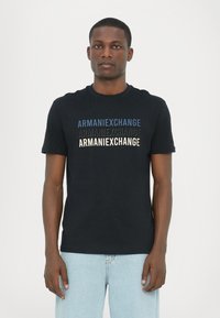 Svart bomull T-skjorte med "ARMANIEXCHANGE"-tekst i blått, svart og kremfarge. Korte ermer og rund hals. Ledig passform.