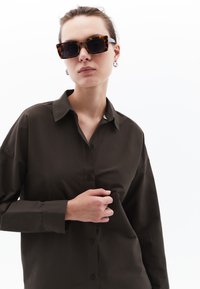 Camicia button-up marrone con colletto classico; presenta maniche lunghe e una vestibilità comoda. Il modello indossa occhiali da sole tartarugati e orecchini d'argento.