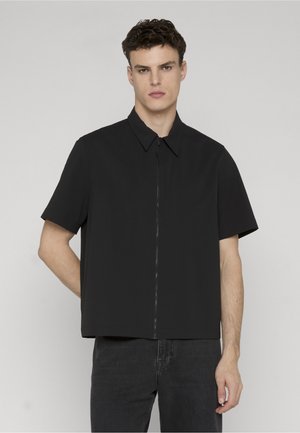 Młody mężczyzna w czarnej koszuli z krótkim rękawem zapinanej na zamek, z kołnierzykiem, stojący na tle jednolitego jasnoszarego tła.