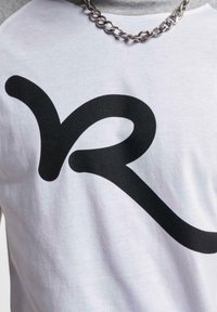 Balta medvilninė marškinė su pilkais raglan rankovėmis, kurių priekyje yra didelis, ryškus juodas "VR" grafikas. Matoma sidabrinė grandinėlė.