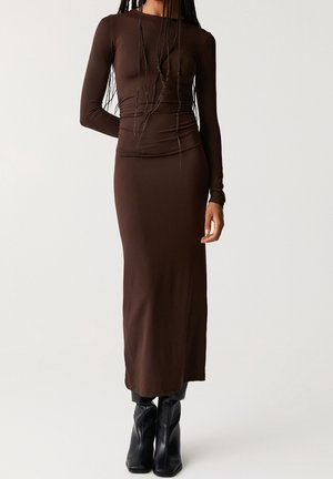 Femme portant une robe midi marron cintrée à manches longues, associée à des bottes noires à bouts pointus, sur un fond blanc uni.