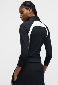 Γυναίκα με πλεξούδες, φορώντας μαύρο και λευκό μπουφάν Nike Shox, με τα χέρια στις τσέπες, κοιτάζοντας πάνω από τον ώμο της σε απλό φόντο.