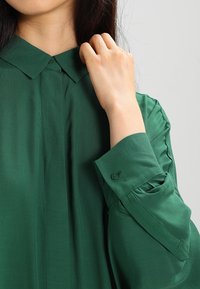 Blusa verde con colletto in tessuto leggero, caratterizzata da dettagli sulla pattina e polsini abbottonati con delicate arricciature sulle maniche.