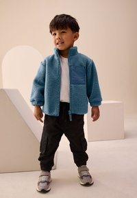 Veste polaire bleue avec fermeture éclair à l'avant, dotée d'une poche poitrine et de poignets côtelés, associée à un pantalon cargo noir et des baskets grises.