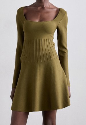 Slim-fit olijfgroene gebreide jurk met lange mouwen, vierkante halslijn en geribbelde details ter hoogte van de taille, gedragen door een staand persoon.