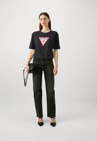 Crna majica kratkih rukava s crvenim trokutastim logom i natpisom "GUESS?". Uparena s crnim trapericama visokog struka i crnim cipelama šiljastih vrhova.