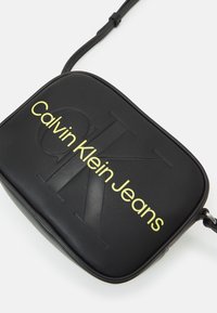 Borsa a tracolla nera in pelle sintetica con logo in rilievo e testo giallo brillante che recita "Calvin Klein Jeans". Include una tracolla regolabile.