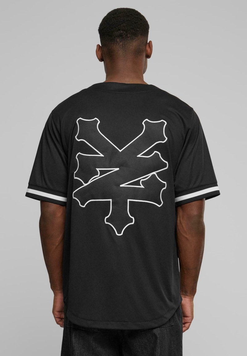 T-shirt noir à manches courtes avec une silhouette blanche d'un crâne et de poignards au dos. Présente des accents rayés sur les manches.