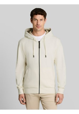 Uomo che indossa una felpa crema con zip e coulisse e pantaloni beige, in piedi davanti a uno sfondo grigio chiaro uniforme.