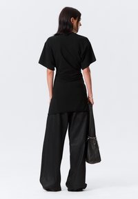 T-shirt preta oversized com mangas curtas e bainha arredondada, combinado com calças largas pretas. O modelo segura uma pequena bolsa preta.