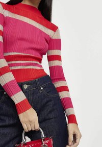 Sweter z prążkowanym wzorem w paski w kolorach różowym, czerwonym i metalicznym, w połączeniu z ciemnoniebieskimi dżinsami. Ręka trzymająca czerwoną torebkę z lakierowanej skóry.