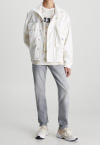 Chaqueta blanca con cremallera y grandes bolsillos, llevada sobre una camiseta de tie-dye claro, combinada con jeans gris claro y zapatillas beige.