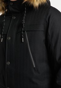 Czarna zimowa kurtka z futrzanym kapturem, pionową kieszenią na zamek, zapięciami na guziki oraz sznurkami z wzorem na fakturowanej tkaninie.