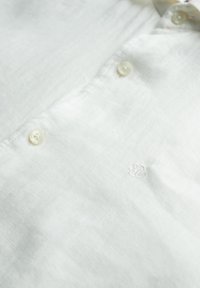 Wit linnen overhemd met een zachte textuur, button-down voorkant en subtiel geborduurd logo aan de linkerkant. De knopen zijn bleek en rond.
