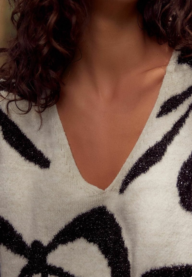 Femme portant un pull blanc duveteux avec des motifs abstraits noirs et un col en V, montrant son cou et ses cheveux bouclés foncés.