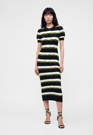 Femme aux longs cheveux noirs portant une robe midi à rayures horizontales noires, blanches et vert olive et des sandales noires à talons hauts.
