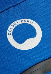 Tejido azul texturizado que presenta un logo circular blanco con el texto "DE LSEY.PARIS", ubicado en el centro. Acentos de goma negra visibles.