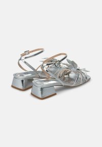 Sandali metallici argento con tacco a blocco, caratterizzati da dettagli floreali e sottili cinturini incrociati alla caviglia, texture liscia e suola piatta.
