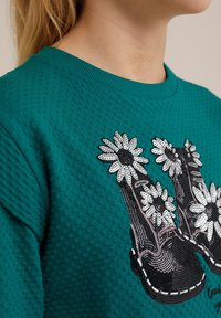Teal színű, textúrázott pulóver, amelyen fekete cipők hímzett mintázata található, fehér özvegyvirágokkal és flitterekkel díszítve. Kerek nyakú.