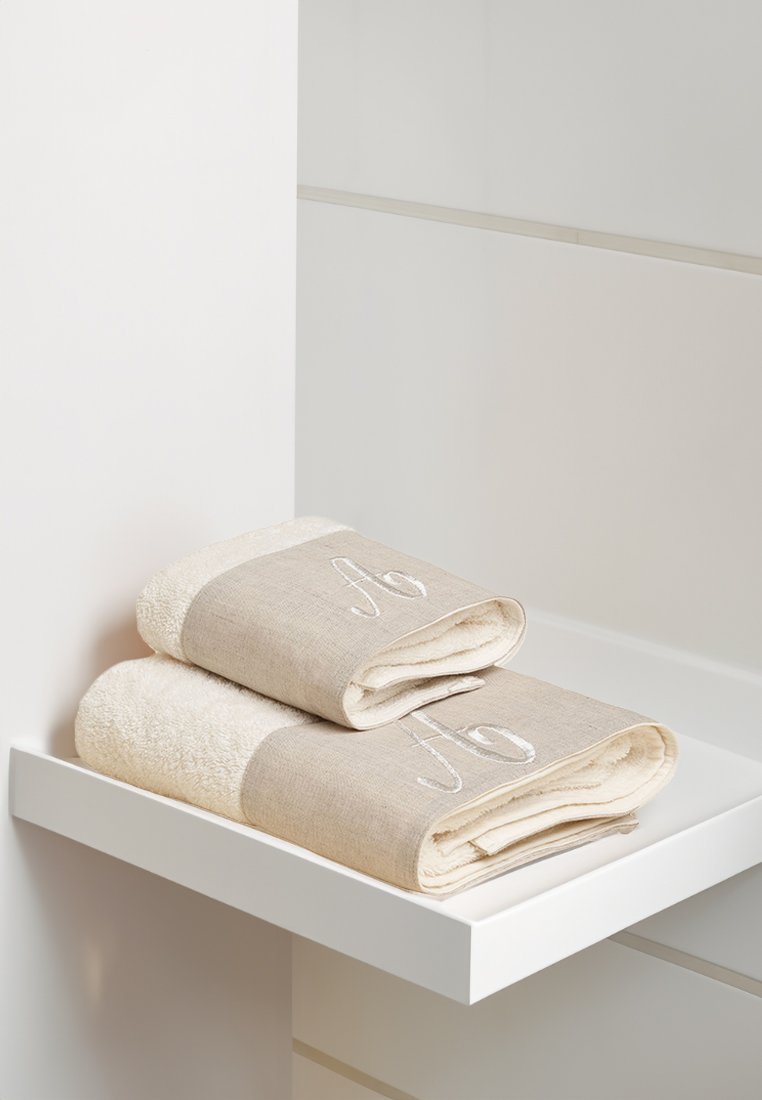 Due stack di due asciugamani poggiati su uno scaffale bianco. Gli asciugamani presentano un tessuto beige con una "A" ricamata sul bordo e un interno soffice bianco.