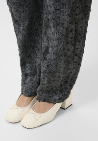 Chaussures à petit talon bloc carré blanches avec de petits nœuds, portées avec un pantalon large texturé sombre.